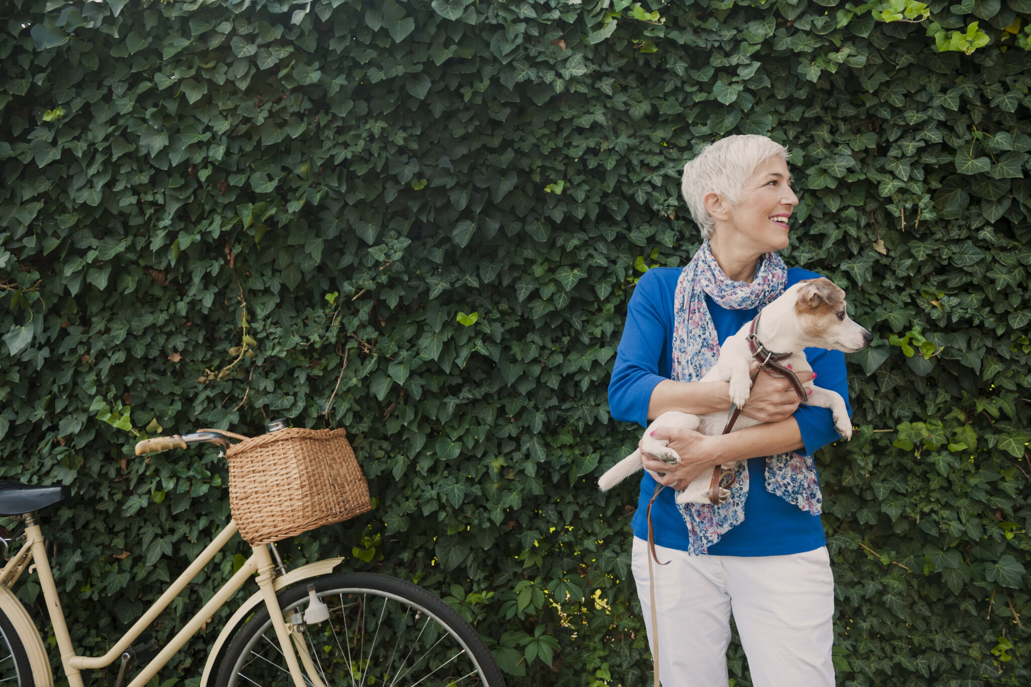 A senior woman holds her beagle in front of an ivy wall, with a bike with a basket leaning against the wall